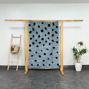 Puede incluir: Una alfombra azul claro con lunares negros se exhibe en un estante de madera. La alfombra tiene una textura peluda y es rectangular. Una pequeña escalera de madera con una cesta de mimbre está a la izquierda de la alfombra.