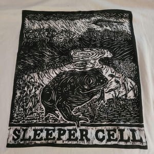 May include: A white t-shirt featuring a black ink-style graphic print of a large toad sitting near a pond in a landscape with trees and rain. The text "SLEEPER CELL" is printed in a bold, blocky font at the bottom of the rectangular illustration.