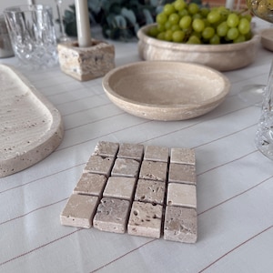 Può includere: Un set di sottobicchieri quadrati in pietra beige su una tovaglia bianca a righe rosse. I sottobicchieri hanno una superficie strutturata. Sono presenti anche una ciotola di uva, un portacandele e bicchieri.