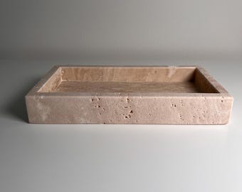 May include: A rectangular, beige stone tray with a recessed interior. The tray has a rough, textured surface and is made of a light-colored stone. The tray is designed to hold small items and is suitable for home decor.