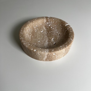 May include: A round, beige stone ashtray with a textured surface and white speckles. The ashtray has a shallow, circular bowl and a thick, rounded base. The neutral color and natural stone texture give it a minimalist aesthetic.