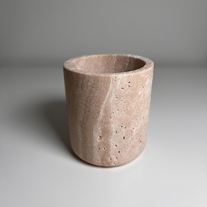 Puede incluir: Una taza cilíndrica de piedra beige con una superficie texturizada. La taza tiene una base redondeada y una parte superior lisa y abierta. La piedra tiene un aspecto natural y poroso, con variaciones de color y textura.