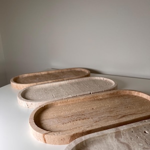 Puede incluir: Cuatro bandejas decorativas ovaladas en tonos beige y marrón. Las bandejas tienen un aspecto texturizado similar a la piedra y están dispuestas en fila. Las bandejas están hechas de un material natural y tienen un borde liso y curvado.
