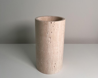 May include: A cylindrical vase made of beige travertine stone. The vase has a smooth, rounded top and a textured surface. The neutral color and simple design make it suitable for various decor styles.
