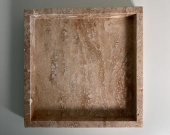 May include: A square, beige travertine tray with a recessed interior. The natural stone features a marbled pattern with variations of light and dark brown. The tray has a simple, modern design, suitable for various uses.