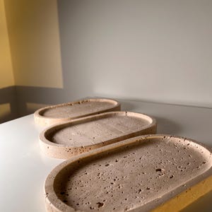 Pode incluir: Três bandejas ovais bege dispostas sobre uma superfície branca. As bandejas têm uma superfície texturizada e porosa e uma borda ligeiramente elevada. As bandejas são feitas de um material semelhante à pedra. Adequado para decoração de casa.