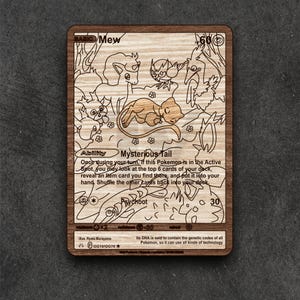 Könnte beinhalten: Holz-Laserschnittkarte mit dem Pokémon Mew. Die Karte hat detaillierte Illustrationen verschiedener Pokémon-Charaktere und Text, der die Fähigkeiten des Charakters beschreibt. Die Karte ist in einem Holzrahmen.