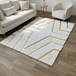 Könnte beinhalten: Ein rechteckiger weißer Teppich mit einem geometrischen Golddesign. Der Teppich hat eine weiche, strukturierte Oberfläche mit goldenen Akzenten, die die Formen umreißen. Der Teppich liegt auf einem Holzboden und ist von Möbeln umgeben.