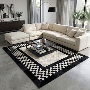 May include: A contemporary living room with a cream-coloured sectional sofa, a black coffee table with books, and a black and white chequered rug. A black floor lamp and a dark wood cabinet complete the scene.
