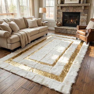 May include: A rectangular white faux fur rug with a gold metallic border. The rug is placed on a hardwood floor in a living room setting, with a sofa, fireplace, and armchair visible in the background.