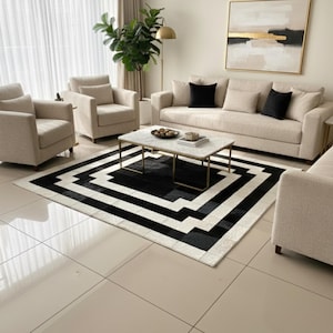 Puede incluir: Un juego de sala moderno con un sofá beige, dos sillones y una alfombra estampada. La alfombra presenta un diseño geométrico en blanco y negro. Una mesa de centro con tapa de mármol se encuentra en el centro y una pintura cuelga de la pared.