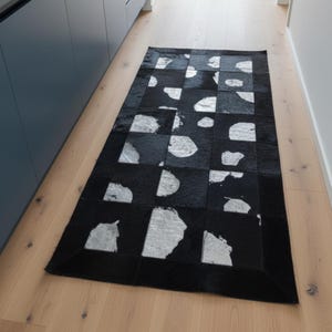 May include: A rectangular black and white cowhide rug with a geometric pattern. The rug features a patchwork design of black squares with irregular white shapes. The rug is placed on a light-coloured wooden floor.