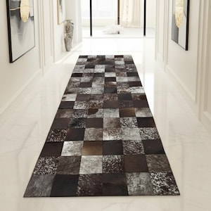 May include: A rectangular hallway rug with a patchwork design. The rug features squares of varying shades of brown, black, and white, creating a textured pattern. The rug is placed in a white hallway with artwork on the walls.