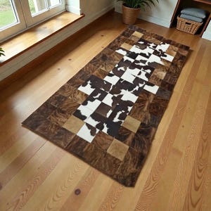 May include: A rectangular cowhide rug with a patchwork design. The rug has a central pattern of alternating black and white squares, surrounded by brown and tan squares. The border is a darker brown. The rug is on a light wood floor.