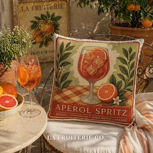 May include: A decorative square pillow with an Aperol Spritz design. The pillow features a large glass of the cocktail with ice cubes and an orange slice, surrounded by leaves and flowers. The text "APEROL SPRITZ" is displayed below the image. The pillow is on a chair.