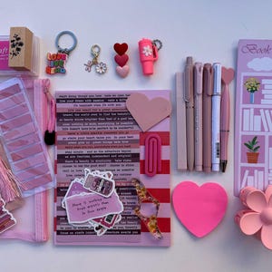 May include: A selection of pink stationery items, including pens, stickers, a stamp, and a notepad. The items are arranged on a white surface, creating a visually appealing assortment for journaling or creative projects.