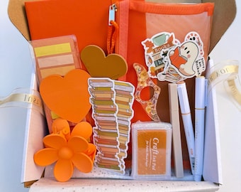 May include: An open cardboard box filled with orange stationery and accessories. Items include a notebook, pencil case, stickers, heart-shaped cutouts, a flower hair clip, pens, and an ink pad. A ribbon with the word "Joy" is visible.