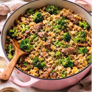 May include: A pink cast iron pan filled with a colourful meal. The dish includes broccoli florets, sliced meat, and grains, garnished with spring onions and sesame seeds. A wooden spoon rests in the pan, and small bowls of spices surround the pan.