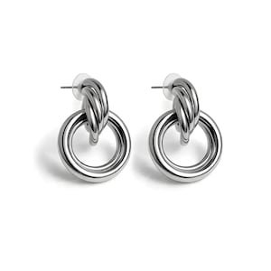 Puede incluir: Un par de pendientes de aro plateados. Cada pendiente presenta un aro grande y circular conectado a un lazo más pequeño y retorcido en la parte superior. Los pendientes tienen un acabado metálico brillante, sobre fondo blanco.