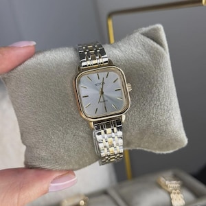 Puede incluir: Un reloj de cuarzo dorado y plateado con esfera cuadrada y correa plateada. La esfera es de color claro con agujas negras y la palabra "NIPON" impresa en la parte superior. El reloj se exhibe sobre un cojín de terciopelo gris.