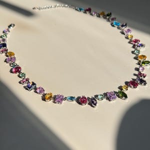 May include: A silver necklace featuring a series of colorful gemstones in various shapes and sizes. The gemstones include pink, purple, yellow, blue, and green hues. The necklace is displayed on a white surface, with sunlight casting shadows.
