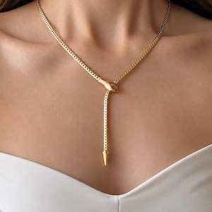 May include: A gold-toned lariat necklace featuring a delicate curb chain. The chain passes through a smooth, elongated metal element shaped like a snake head, with a pointed, tapered metal tip hanging down at the end of the drop section.