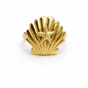 Puede incluir: Anillo dorado con diseño de concha y estrella de mar. El anillo tiene un acabado pulido, con la concha formando el cuerpo principal y la estrella de mar centrada en la parte delantera. El anillo está sobre un fondo blanco.