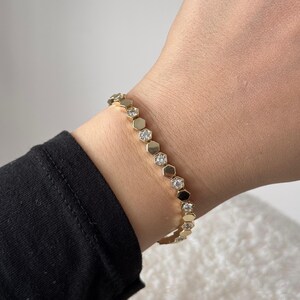 May include: A gold bracelet with a honeycomb design, featuring alternating hexagonal links. Some links are adorned with clear, sparkling gemstones. The bracelet is worn on a wrist, with a black sleeve visible.
