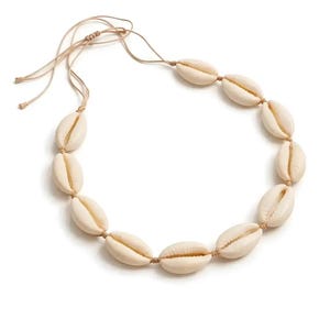 Puede incluir: Un collar con una serie de conchas de cauri de color crema ensartadas en un cordón beige. Las conchas son ovaladas y tienen una textura natural y suave. El cordón es ajustable.