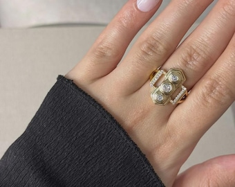 Könnte beinhalten: Ein goldener Ring im Art-Deco-Stil mit drei zentralen runden Diamanten und kleineren Akzentsteinen. Der Ring befindet sich an einer Hand mit langen, weiß getippten Nägeln, und die Person trägt einen schwarzen Ärmel.