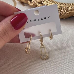 May include: Gold-toned jewelry set featuring three pairs of earrings. One pair is a small hoop, another is a safety pin design, and the third is a small drop earring. The earrings are embellished with small, clear stones. The word "JEWELRY" is printed on the card.