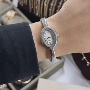 Puede incluir: Reloj ovalado plateado con esfera blanca y números romanos negros. El reloj tiene un movimiento de cuarzo y una pulsera de estilo brazalete plateada. El reloj se lleva en la muñeca, con una manga negra visible.