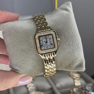 May include: Gold-coloured wristwatch with a rectangular face and a gold link band. The watch face is white with black Roman numerals and hands. The bezel is encrusted with small, clear crystals. The watch is displayed on a grey pillow.