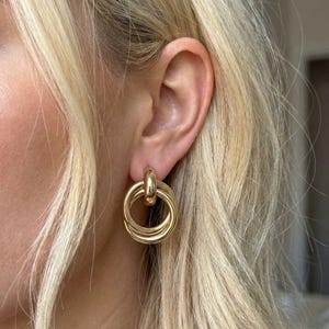 Könnte beinhalten: Goldfarbene Creolen mit einem dreifachen Kreisdesign. Die Ohrringe sind an einem Stecker befestigt und werden an einem Ohr mit blondem Haar im Hintergrund gezeigt. Die Ohrringe haben eine polierte Oberfläche und einen klassischen, eleganten Stil.