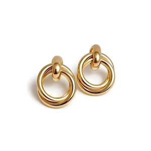 Puede incluir: Un par de pendientes dorados. Cada pendiente presenta un diseño de doble bucle con una barra de conexión en la parte superior. Los pendientes tienen una superficie pulida y reflectante, sobre un fondo blanco.