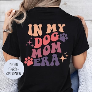 May include: Black t-shirt with the text "IN MY DOG MOM ERA" in retro-style lettering. The design includes paw prints and stars in shades of pink, orange, and purple. A round white sticker with German text is on the left.