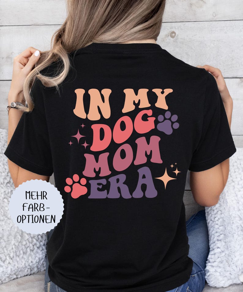 May include: Black t-shirt with the text "IN MY DOG MOM ERA" in retro-style lettering. The design includes paw prints and stars in shades of pink, orange, and purple. A round white sticker with German text is on the left.
