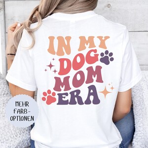 May include: White t-shirt with the text "IN MY DOG MOM ERA" in orange, red, and purple. The design includes paw prints and starbursts. The shirt is a casual style.
