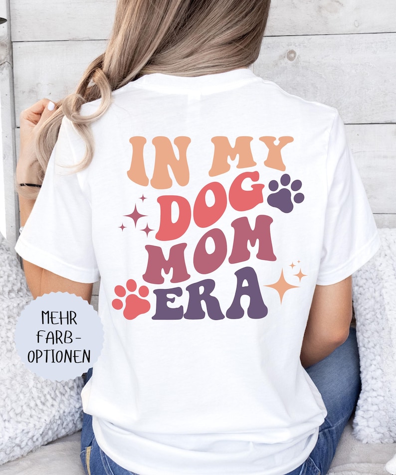 May include: White t-shirt with the text "IN MY DOG MOM ERA" in orange, red, and purple. The design includes paw prints and starbursts. The shirt is a casual style.