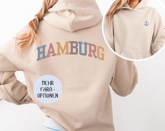 May include: A beige hooded sweatshirt featuring the word HAMBURG in a multi-coloured arched font on the back. A circular inset shows the front of the same sweatshirt with a small blue anchor graphic on the chest and a light blue circular badge.