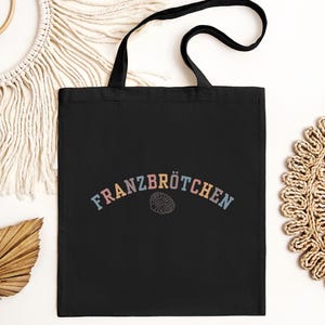 May include: Black tote bag featuring the word "FRANZBR&Ouml;TCHEN" in pastel colours, arched across the top. Below the text is a line drawing of a pastry. The bag has black handles and is set against a neutral background.