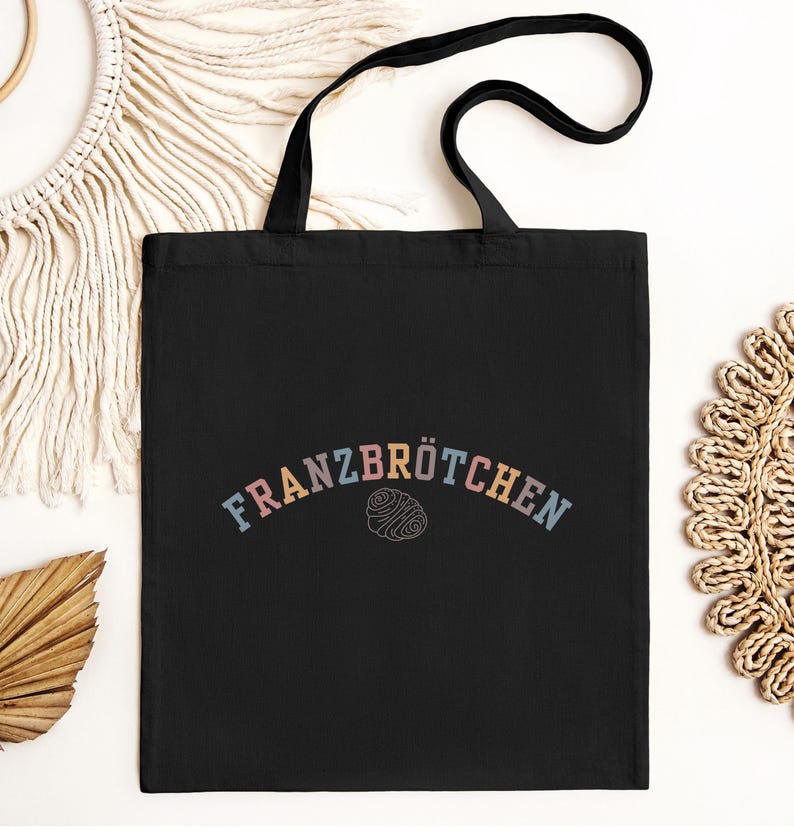 May include: Black tote bag featuring the word "FRANZBR&Ouml;TCHEN" in pastel colours, arched across the top. Below the text is a line drawing of a pastry. The bag has black handles and is set against a neutral background.