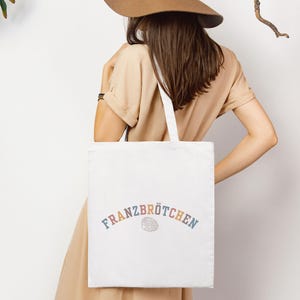 May include: A white tote bag featuring the word "Franzbr&ouml;tchen" in rainbow lettering. The bag is carried by a person wearing a tan dress and a brown hat. The bag's design is simple, suitable for shopping or everyday use.