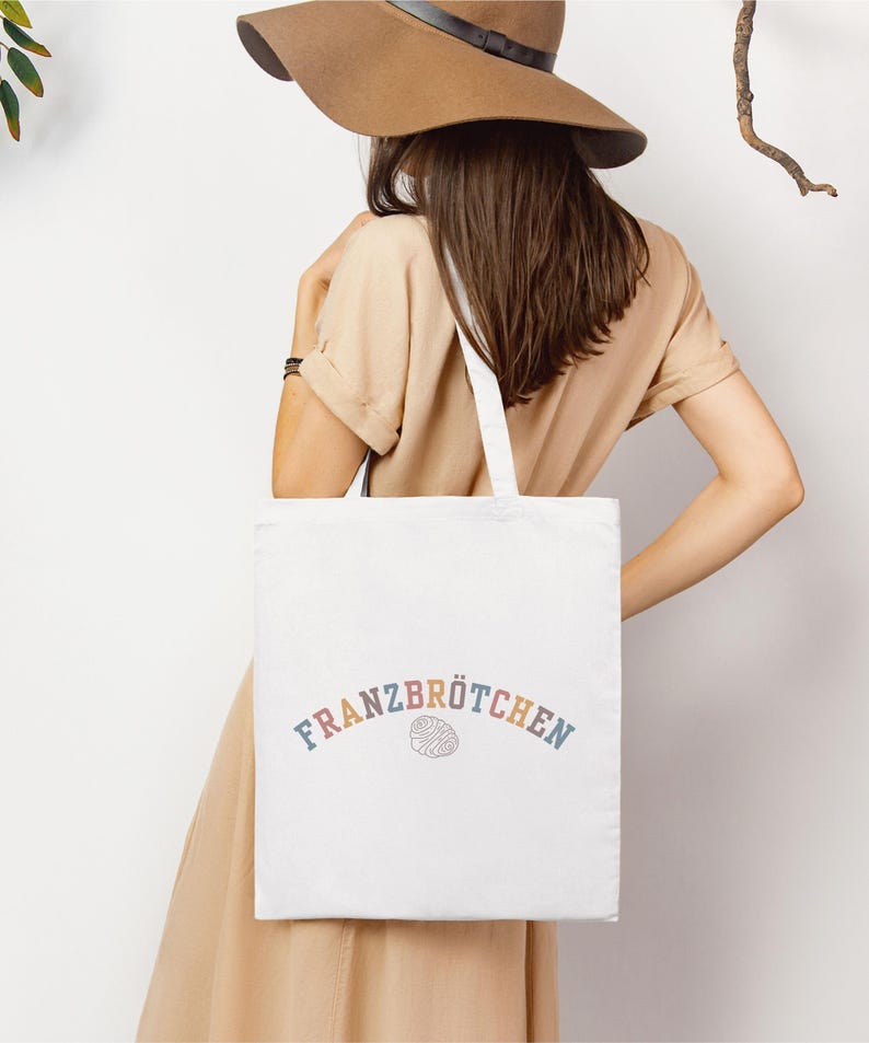 May include: A white tote bag featuring the word "Franzbr&ouml;tchen" in rainbow lettering. The bag is carried by a person wearing a tan dress and a brown hat. The bag's design is simple, suitable for shopping or everyday use.