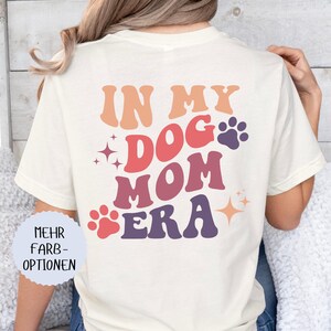 May include: Cream-colored t-shirt with the text "IN MY DOG MOM ERA" in retro-style lettering. The design includes paw prints and starburst accents in shades of orange, red, and purple. The shirt is a casual style.