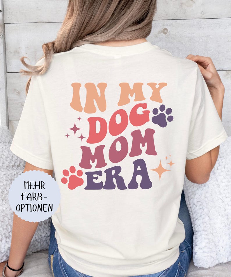 May include: Cream-colored t-shirt with the text "IN MY DOG MOM ERA" in retro-style lettering. The design includes paw prints and starburst accents in shades of orange, red, and purple. The shirt is a casual style.