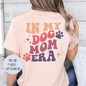 May include: A light peach-colored t-shirt with the words "IN MY DOG MOM ERA" in a retro font. The design includes paw prints and star accents. The shirt is a comfortable, casual style.