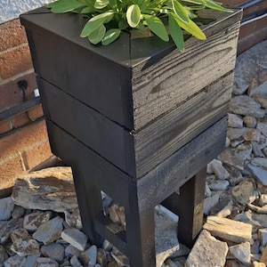 May include: A dark stained wooden planter, approximately 40 cm high, filled with green foliage. The planter has a square top and is supported by four legs. It rests on a bed of grey and brown stones, with a white wall and brickwork in the background.