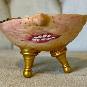 Puede incluir: Un cuenco caprichoso hecho a mano con un diseño de rostro. El cuenco está sostenido por tres patas doradas. El exterior del cuenco está pintado con un color carne, con una nariz, boca y dientes. La boca está pintada con detalles rojos y blancos.