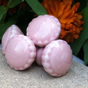 May include: Four pink ceramic cabinet knobs with a scalloped edge. The knobs are arranged in a cluster on a light grey surface.